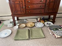 Assorted plates, bowls, trays, linens, and serving ware displayed on carpet in front of wooden cabinet, showing a variety of items including floral ceramic plates, clear glass trays, and folded linens.