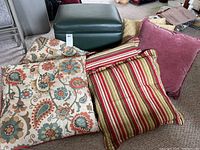 Photo showing green leather storage ottoman on wheels with six decorative pillows of various patterns and textures arranged on the floor.