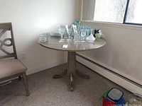 Round wooden table positioned in corner by windows, showing clear tabletop with some glassware and items on top for space context.