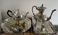 Two silverplate teapots on silverplate serving platters with serving utensils and salt cellar