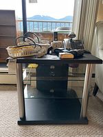Front view of a black and silver TV stand with glass shelves, showing top surface with miscellaneous items that are not included.