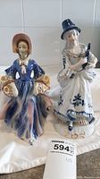 Pair of two vintage porcelain figurines, one in blue dress with flower baskets and one in white dress with blue patterns and bonnet, placed on white cloth.