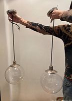 Two clear globe pendant hanging lamps shown held by a person, featuring metal canopy tops, black cords, and large clear spherical glass shades.