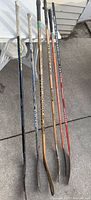 Full view of six hockey sticks leaning against a wall showing length and grip condition.