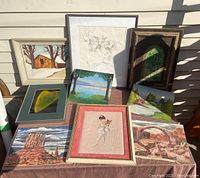 Overview of nine paintings and sketches arranged on a table outdoors in sunlight.