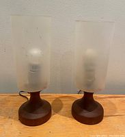 Pair of MCM table lamps with solid teak bases and frosted plastic shades on a wooden surface, showing front view.