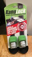 Front view of KanuLock utility straps in original packaging with two green lockable buckle covers and black straps, showing use case on roof racks with kayak.
