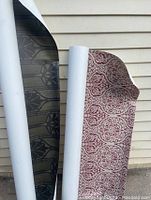 Photo showing two large wallpaper rolls leaning against an exterior wall. One roll shows burgundy damask pattern, the other black floral geometric pattern.