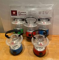 Lot of four LED camping lanterns still in original packaging, showing the red, blue and green colored bases with black handles and translucent tops.