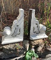 Pair of Victorian architectural corbels positioned outdoors on rocks, showing side views with curved scroll details and cutout patterns.