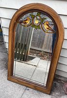 Full view of arched wooden framed mirror standing outside showing stained glass decoration on top half and wood grain frame