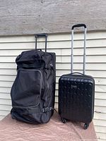 Front view of two pieces of luggage: black Roots soft rolling duffel on left and black Samsonite hard shell rolling suitcase on right against beige siding