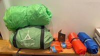 Overall lot with green tent packs, orange and blue straps, sleeping pads and manual arranged on bench