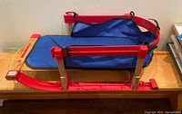 Side and top angled view of child's sleigh showing wooden frame, blue fabric seat, red metal runners, and attached yellow rope.