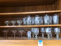 Shelf with 15 clear drinking glasses with floral design and pink-tinted champagne coupe glasses.
