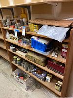 Wide shot of wooden shelving unit holding jars, boxes, and containers filled with various nails, screws, and other hardware items. Also includes a metal toolbox and miscellaneous small parts.