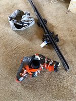 Photo of black and orange ski boots placed on carpet floor near black skis with bindings and grey ski bag.