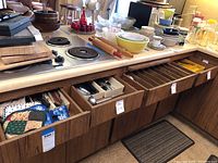 Wide view of open drawers containing various cutlery and silverware with kitchen countertop items including bowls, jars, and rolling pins.
