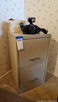 Beige two drawer metal file cabinet standing upright with a camera and white container placed on top. Shows overall front view and condition.