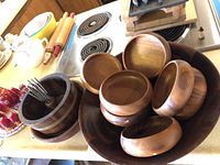 Photo showing several wooden bowls stacked on a countertop with rolling pins and other kitchen items in the background.