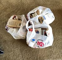 Three plastic grocery bags on carpet, each filled with multiple paperback books visible throughout openings.