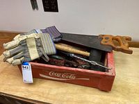 Photo showing work gloves, handsaw, mallets, and other tools stored in Coca-Cola crate on wooden table.
