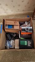 Top view of cardboard box containing various cameras and accessories including Sears Super 8 movie camera and Olympus camera kit
