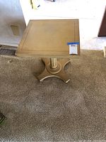 Square wooden end table with a light beige finish shown on carpeted floor, scratches visible on the tabletop surface