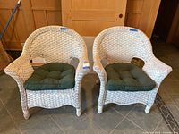 Pair of white wicker chairs with green cushions sitting on a tile floor, showing both chairs from front angle.