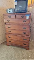 Front view of the wooden chest of drawers showing five large drawers and two small top drawers, items on top include a TV and radio.