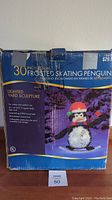 Photo of original box showing the 30 inch frosted skating penguin lighted yard sculpture with details including lighted yard sculpture suitable for indoor and outdoor use, long-lasting bulbs, collapsible construction.