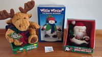 Photo of three Christmas plush toys: a moose plush, boxed dancing snowman plush, and boxed Santa Claus plush.