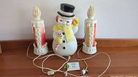 Full view of the three vintage Empire blow mold decorations: one snowman and two Noel candles with cords for illumination on a wooden surface against a white wall