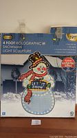 Front view of the closed box showing the image of the holographic snowman light sculpture, product details, and retail price of $19.99.