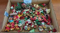 Box filled with assorted vintage Christmas ornaments including Santas, snowmen, reindeer, candy canes, bells, and greenery with pine cones.