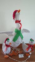 View of three lighted Christmas geese yard decorations showing largest goose with green scarf and two smaller geese with red bows and tinsel feathers