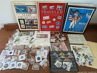 Overall view of the collection including three stamp albums, loose coins in sleeves, stamps in pages, paper money, pins, and other collectibles laid out on a wooden surface.