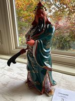 Side view of the blue-green robed Guan Yu figurine holding a curved weapon, with detailed gold embellishments and standing on patterned shoes.