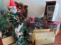Photo showing several Christmas wreaths, inflatable Santa Claus figure, boxes of Kirkland holiday decorations and Christmas lights, and a box with red shiny ornaments.