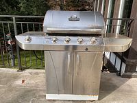 Front view showing the full stainless steel BBQ grill with four control knobs and two side shelves, rust visible on lower edges.