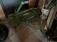 Green metal tiller attachment with bar handle and curved tines, placed on a floor inside a barn or shed.