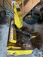 Side angle view of yellow John Deere tractor snowblower attachment showing hydraulic hoses and yellow body with rust and paint wear.