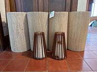 Front view of three large beige textured cylindrical lamp shades and two smaller teak wood framed lamp shades with fabric lining.