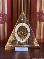 Front view of Seiko triangular mantel clock showing clear triangular body with brass accents and exposed clock mechanism with Roman numeral dial.