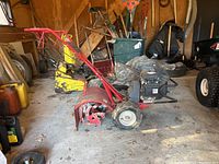 Side overview of red Troy-Bilt tiller on garage floor showing tines and handle