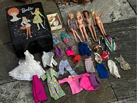 Overview of four dolls laid out with assorted vintage doll clothes and shoes around them, plus the Barbie carrying case.