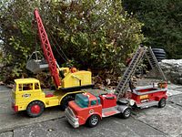 Side view of the yellow crane truck with red crane arm and the red fire truck with trailer ladder parked outdoors on stone surface.