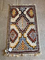 Full view of the small rectangular carpet with geometric pattern in burgundy, yellow, blue, black on cream background, laid flat on floor.