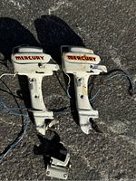 Pair of white vintage Mercury electric toy boat motors with red Mercury logo, showing front view and side by side arrangement on pavement surface.
