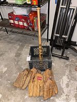 Photo showing the wood bat standing in a crate with five leather gloves arranged in front on concrete floor.